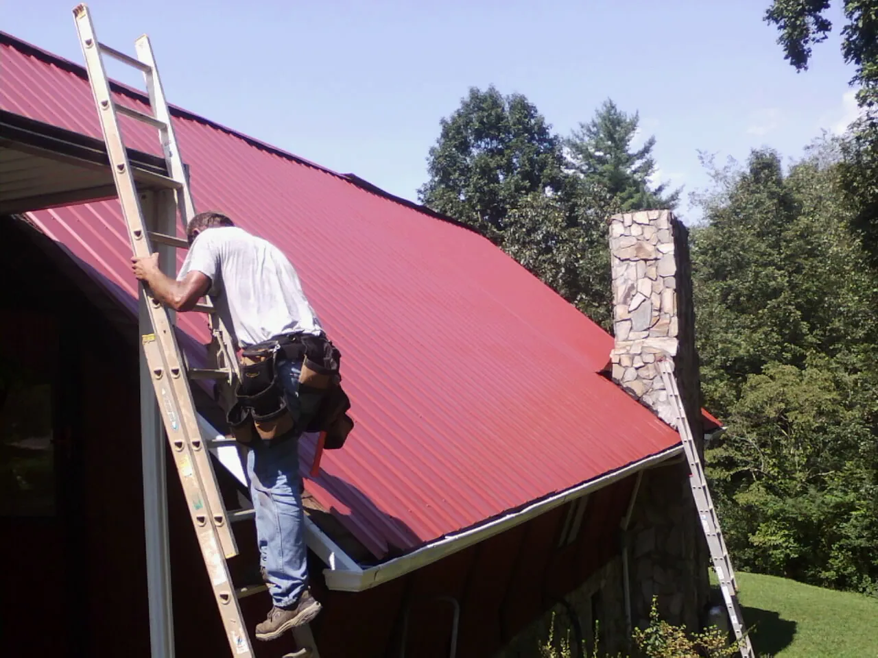 Roofer on a ladder beside a red metal roof and stone chimney — Soffit & Fascia Repair by Roofing Brothers in Summitville